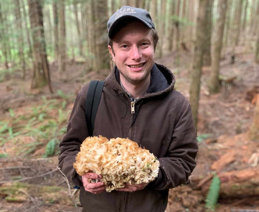 Color photo of Bark's Forest Watch Coordinator, Michael Krochta, holding a cauliflower mushroom from Bark's 2019 mushroom hike. He's wearing a brown hoodie and gray cap and smiling at the camera.