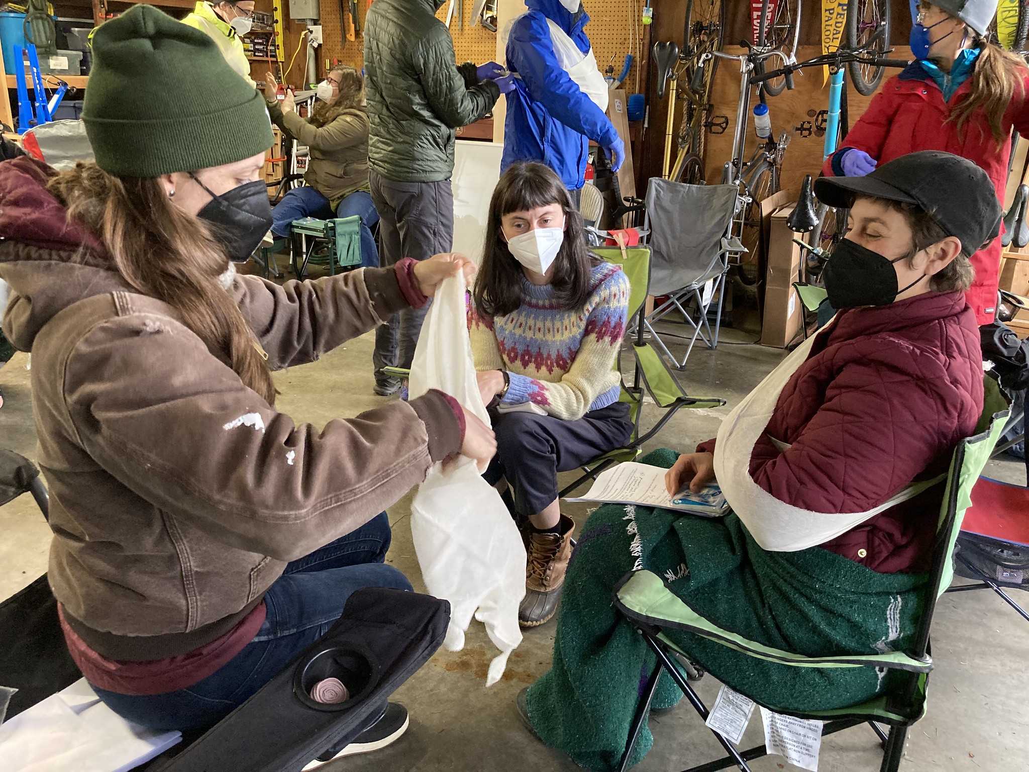 This image shows Daniela, a volunteer, and a member of the Rose Hips Forest Aid group demonstrating how to apply a sling for a hurt arm. This group is gathered at an indoor garage setting.