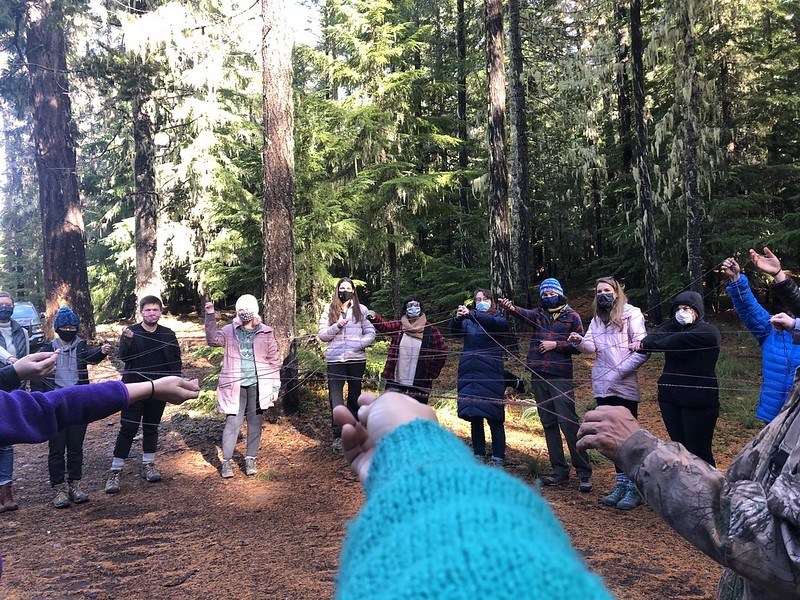 Image shows a group of staff and volunteers gathered in a forest clearing, connected to one another by a large web of string.