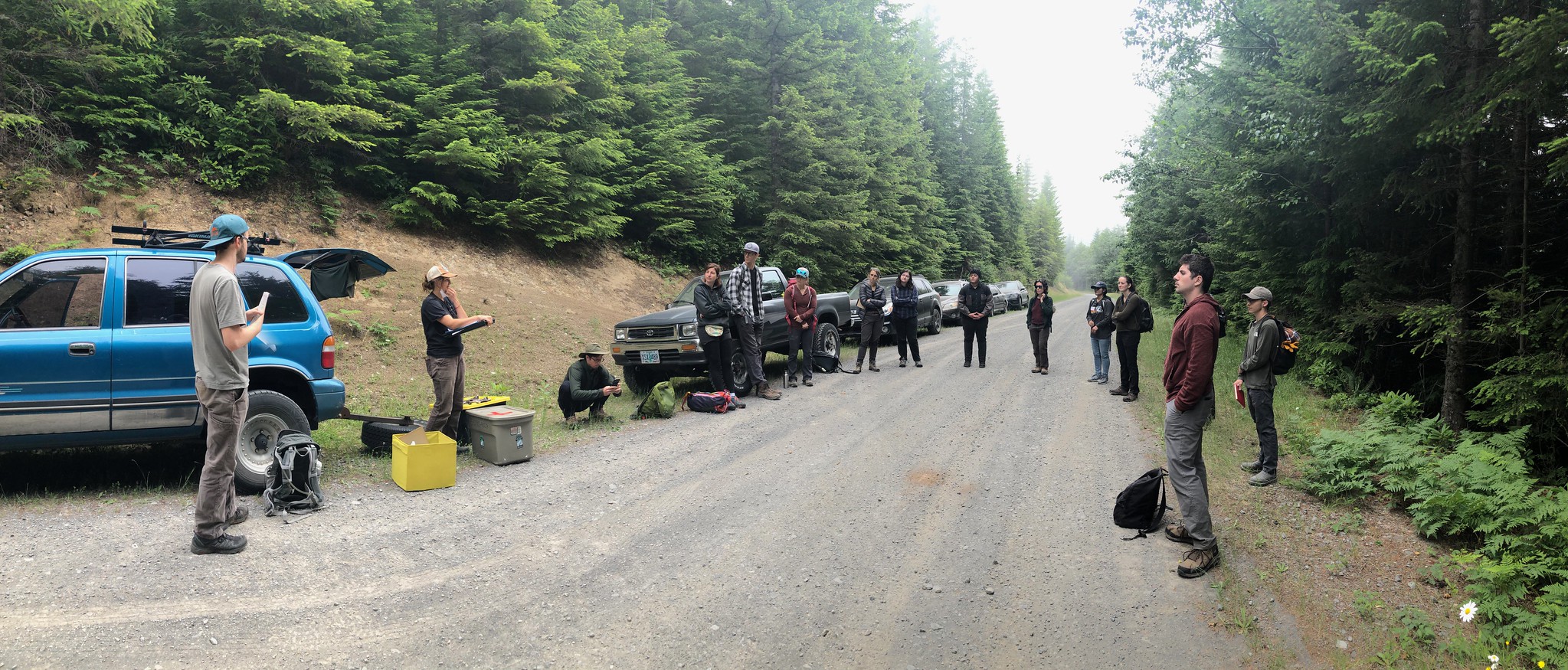 A group of 14 people standing in a semi-circle on a gravel road with vehicles parked behind them.