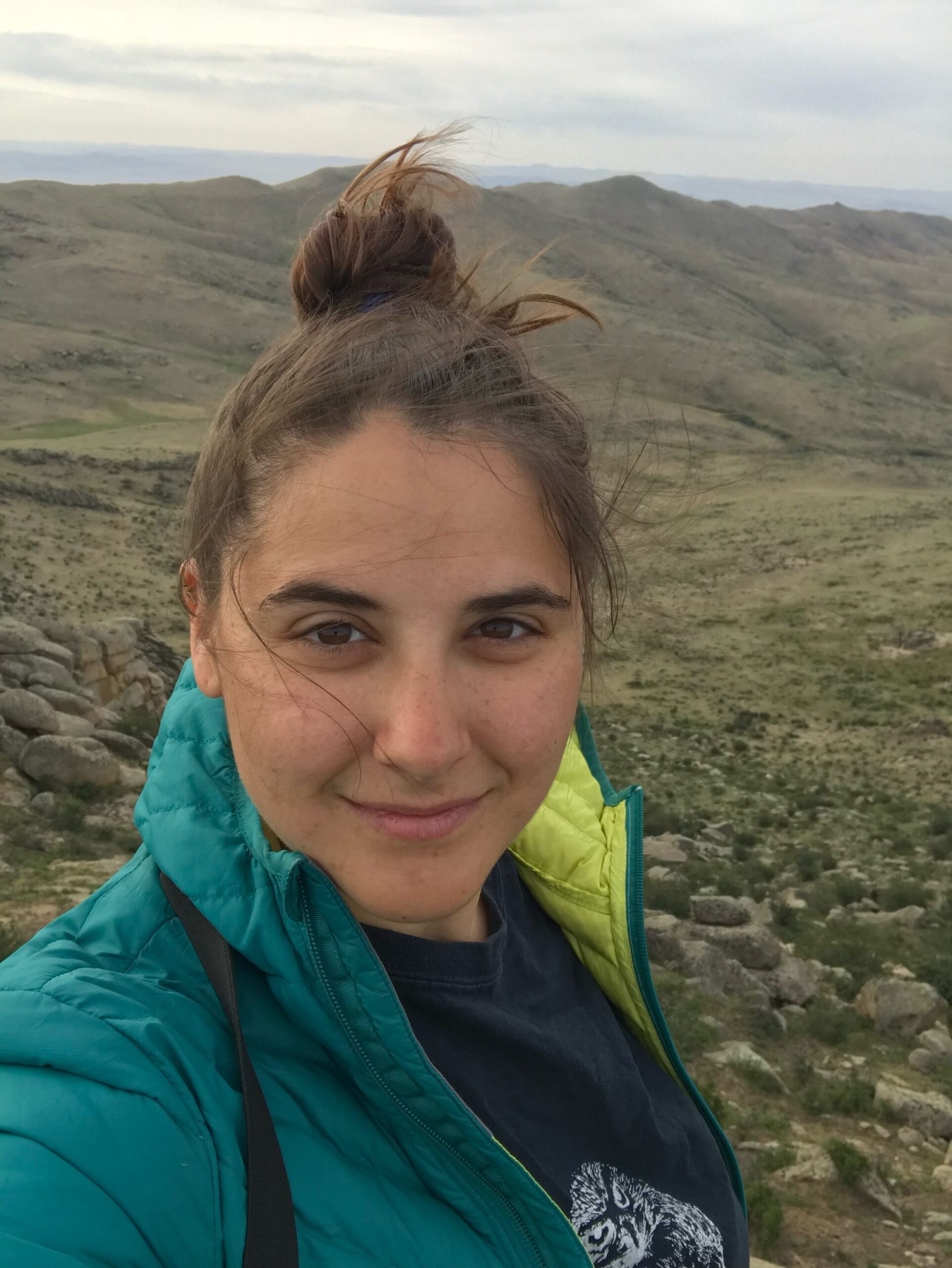 This image shows Christine in a rocky desert landscape, wearing a blue jacket and with her brown hair up in a bun and a soft smile on her face.