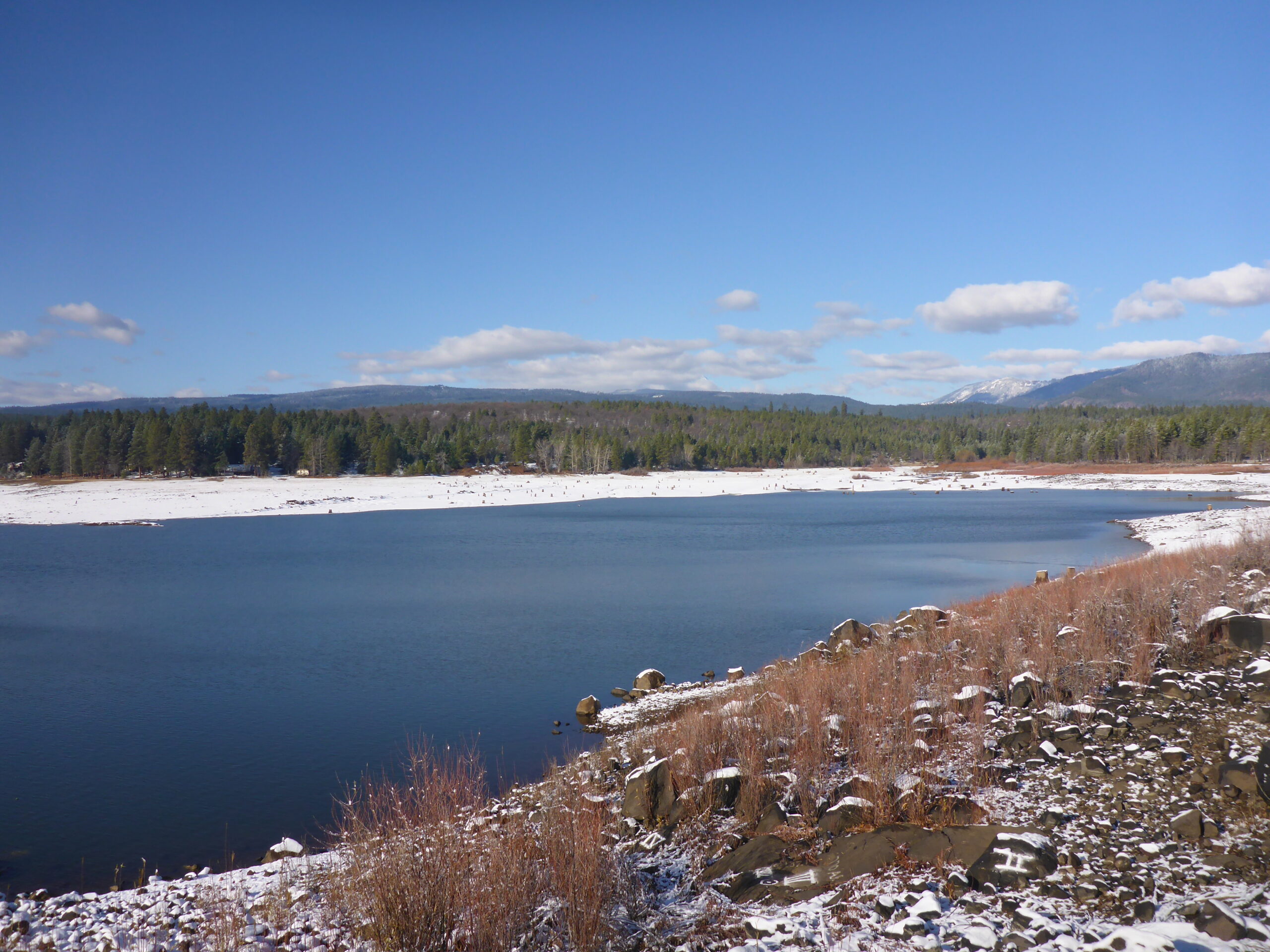 This is an wideview of Rock Creek Reservoir with the Rocky Timber Sale in the background. The shores of the reservoir is covered in snow and the sky is a deep blue color.
