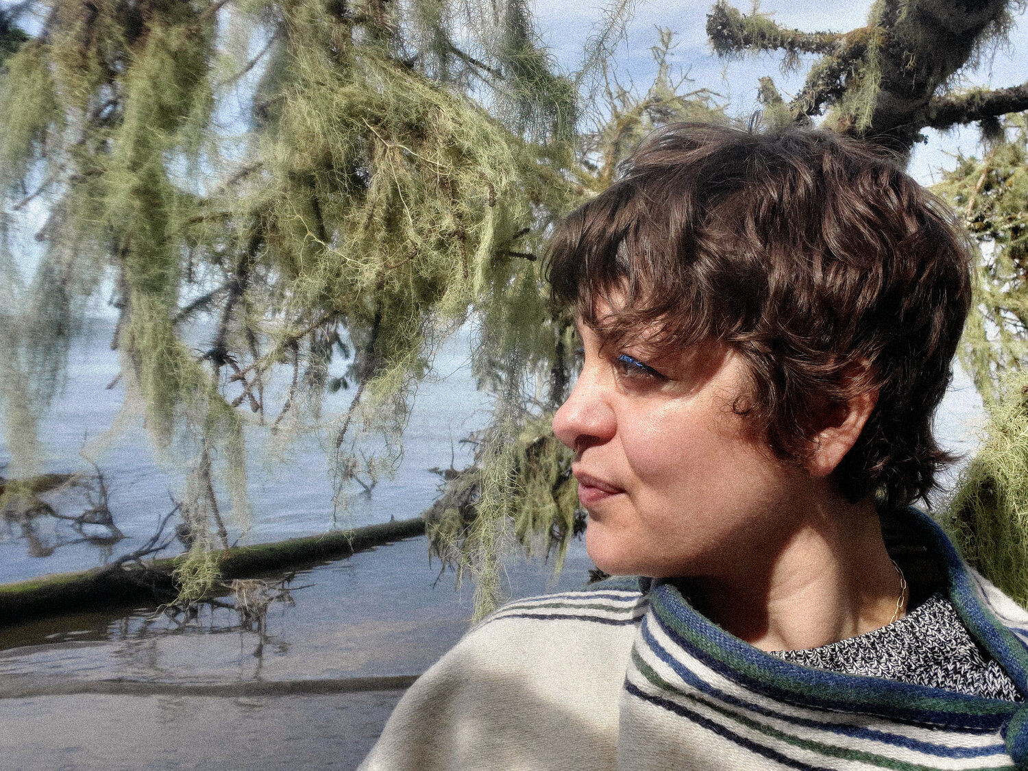 Headshot of Daniela del Mar wearing a striped poncho in front of lichen off the coast of Astoria
