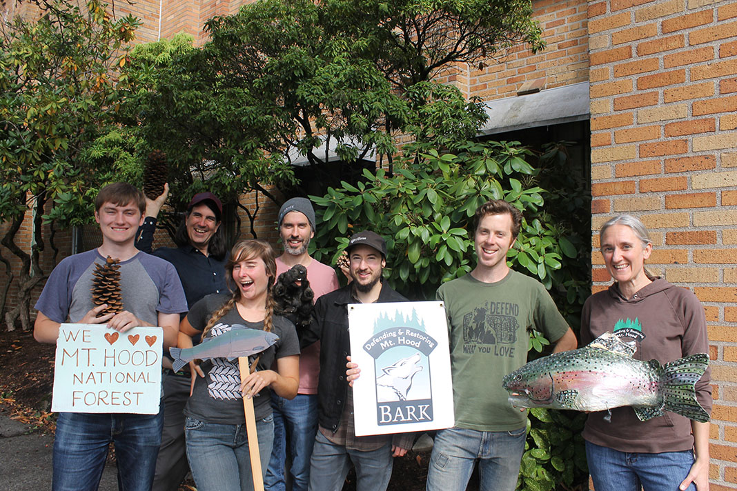 Color photo of a group of people by the Bark office, in front of a rhodedendron and brick wall holding Bark ephemera smiling for the camera