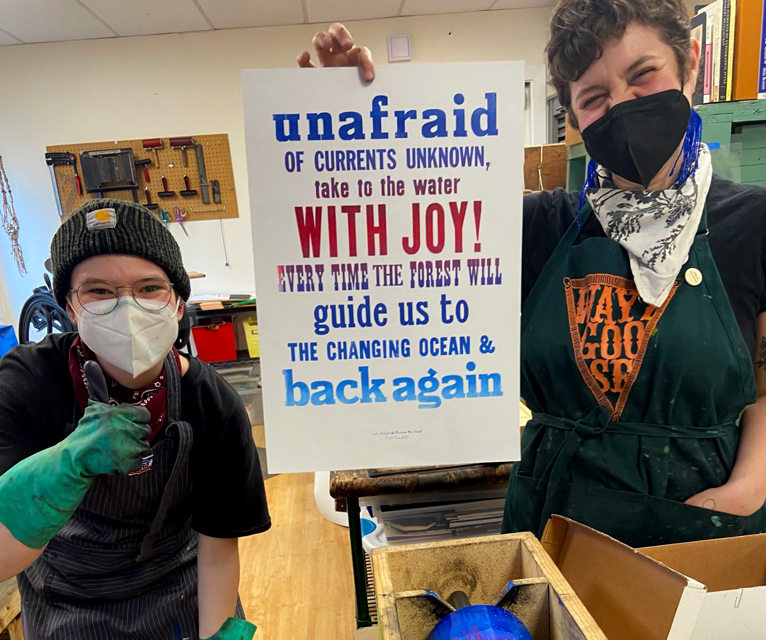 [Two nonbinary individuals (one with a beanie and one with brown hair) are facing the camera and wearing dark clothing, aprons, and gloves. They are holding up a poster printed in a gradient that reads 